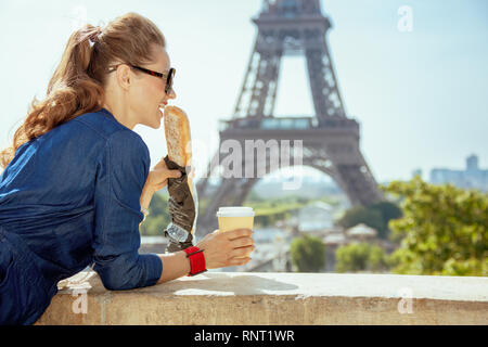 Voyageur élégant smiling woman in blue jeans avec global Coffee cup baguette manger avoir entière contre vue claire de la Tour Eiffel à Paris Banque D'Images