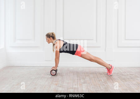 Vue côté profil portrait de jeune femme blonde athlétique en fit porter à l'aide de rouleau en mousse dans une salle de sport à la séance d'entraînement au poids de perte et faire plank. Rehabilitati Banque D'Images