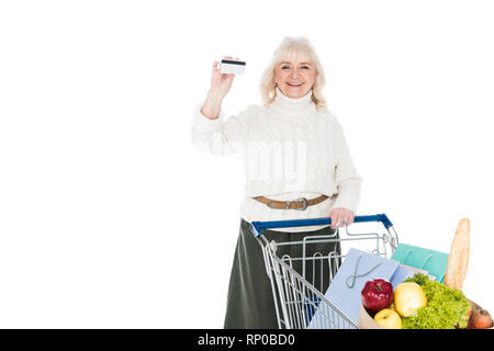 Smiling senior woman holding credit card near shopping trolley isolated on white Banque D'Images