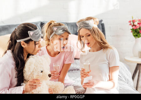 Belle surprise in multiculturelle masques couchage avec verres de champagne en utilisant smartphone pendant soirée pyjama Banque D'Images