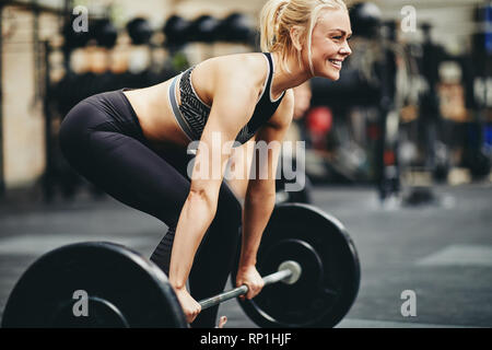 Fit young woman smiling in sportswear tout en se préparant à lever des poids lourds au cours de l'exercice dans la salle de sport Banque D'Images