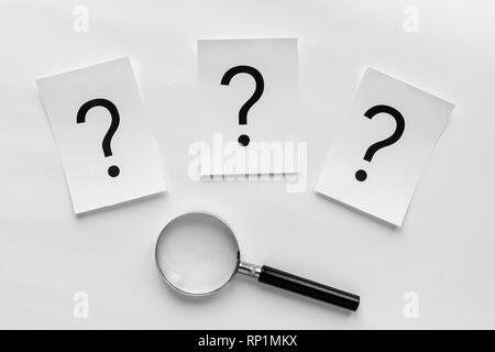 Trois points d'interrogation imprimés sur cartes blanches dans une ligne au-dessus d'une loupe sur un fond blanc dans une image conceptuelle Banque D'Images