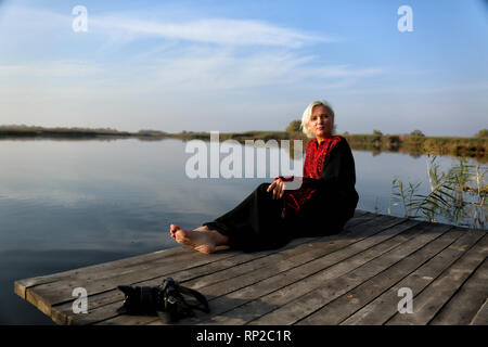 Au printemps, une belle femme dans une robe brodée de bourgogne repose sur une jetée en bois près de l'eau calme avec un appareil photo dans un beau temps. Banque D'Images