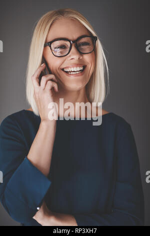 Young businesswoman laughing et parler sur son portable tout en se tenant sur un arrière-plan gris Banque D'Images