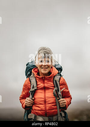 Smiling woman hiker portant veste et sac à dos sur une randonnée. La recherche d'aventure femme marche sur une colline sur un jour nuageux. Banque D'Images