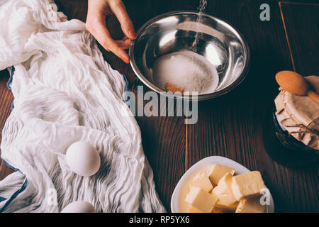 Close-up woman cooking pâte à tarte verser sur le sucre d'œuf dans un bol en métal à côté d'oeufs, le beurre coupé en dés et pot de confiture, vue du dessus sur table en bois brun. Banque D'Images