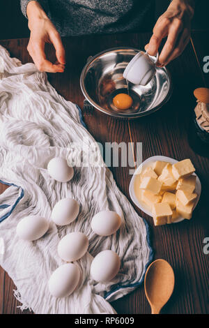 Préparer la pâte à tarte Femme versant d'œuf dans un bol en métal à côté d'oeufs et le beurre coupé en dés, vue du dessus sur table en bois brun. Banque D'Images