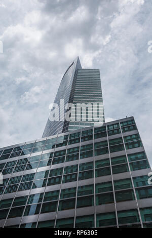 Une prise de vue sous-jacente capture la grandeur architecturale de deux gratte-ciels modernes sur fond de ciel nuageux. Le plus grand bâtiment, avec sa façade en verre épurée, se rétrécit jusqu'à un point en haut, tandis que la structure inférieure présente un motif de fenêtres en forme de grille, reflétant le paysage urbain environnant. Banque D'Images