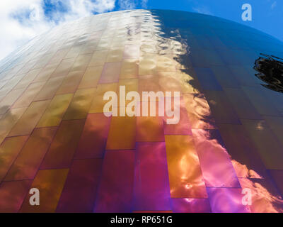 Une prise de vue en angle bas capture l'extérieur du Musée de la culture pop (MoPOP) à Seattle, Washington, mettant en valeur son design architectural unique. La façade du bâtiment est composée de panneaux métalliques irisés qui reflètent le ciel et les nuages, créant un effet dynamique et visuellement saisissant. Les panneaux passent des teintes or et orange en bas aux tons bleu et argent en haut, reflétant le dégradé du ciel. Ce design vise à incarner l’énergie et la fluidité de la culture pop, faisant du MoPOP une destination phare pour les visiteurs. Banque D'Images