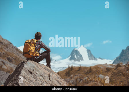 Homme hiker with backpack est assis sur un gros rocher et s'intéresse à la montagne blanche avec glacier Banque D'Images