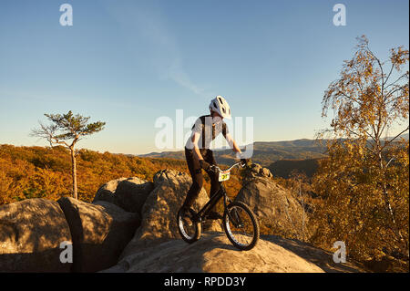 Les jeunes cyclistes masculins en équilibre sur le vélo d'essai haut de big boulder, rider stunt acrobatique faire sur soir d'été, ciel bleu et de la forêt sur l'arrière-plan. Concept de sport extrême de vie actif Banque D'Images