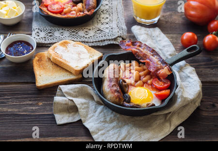 Les œufs, saucisses, bacon, haricots et champignons dans la poêle de fer, toasts, beurre, jus d'orange sur fond de bois rustique. Le petit déjeuner anglais complet. Banque D'Images