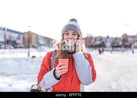 Happy Girl in red winter jacket fait appel téléphonique. Happy smiling in hand tasse café thé chaud. Les émotions de joie et de plaisir, de plaisanterie drôle Banque D'Images