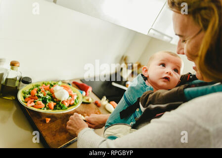 Smiling baby porté dans un porte-bébé sac à dos à la recherche de sa mère alors qu'elle cuisine, concept de conciliation familiale Banque D'Images