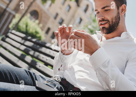 Bel homme va faire appel à l'aide d'écouteurs. Homme barbu tenant des écouteurs, des vêtements décontractés, chemise blanche, jeans, attrayant, homme d'affaires face au parc. Portrait de jeune homme à l'extérieur, élégant costume. Guy avec écouteurs et smartphone assis sur un banc. Banque D'Images