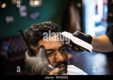 Jeune homme barbu se coupe par coiffure avec rasoir électrique tout en restant assis dans fauteuil de barbier Banque D'Images