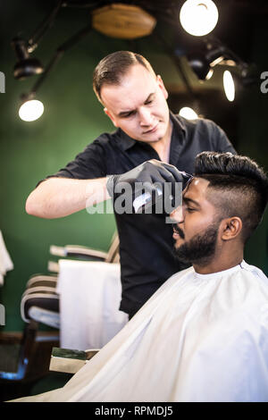 Jeune homme barbu se coupe par coiffure avec rasoir électrique tout en restant assis dans fauteuil de barbier Banque D'Images