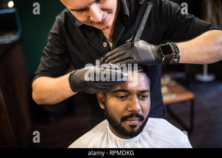 Jeune homme barbu se coupe par coiffure avec rasoir électrique tout en restant assis dans fauteuil de barbier Banque D'Images