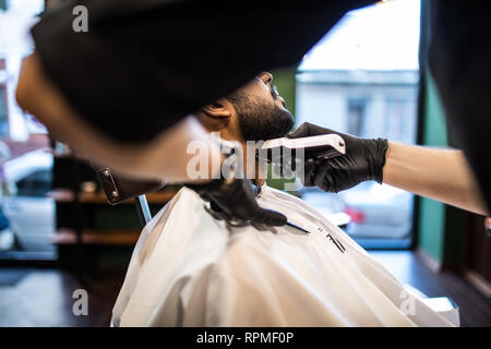 Le toilettage de l'homme réel. Vue latérale du jeune homme barbu se coupe de barbe à coiffure while sitting in chair at barbershop Banque D'Images