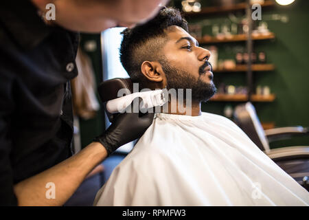 Jeune homme barbu se coupe par coiffure avec rasoir électrique tout en restant assis dans fauteuil de barbier Banque D'Images