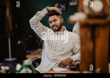 Temps pour nouvelle coupe. Beau barbu en regardant son reflet dans le miroir et en gardant la main dans les cheveux tout en sitting in chair at barbershop Banque D'Images