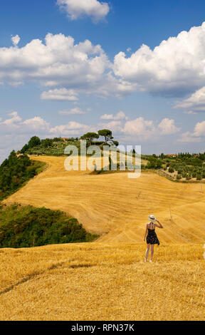 Une jeune femme avec une robe courte et un chapeau de paille d'admirer les collines en face d'elle à partir d'un champ de foin coupé dans le Val d'Orcia, Toscane, Italie Banque D'Images