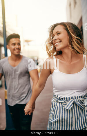 Souriante jeune femme menant son petit ami par la main tout en marchant ensemble dans une rue de la ville Banque D'Images