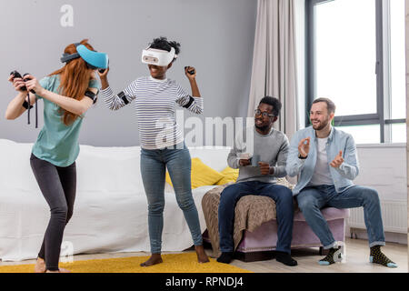 Deux jeunes femmes multiraciales portant des lunettes de réalité virtuelle de danse à la maison pendant que leurs petits amis assis sur le canapé et souriant, profitant de l'action Banque D'Images