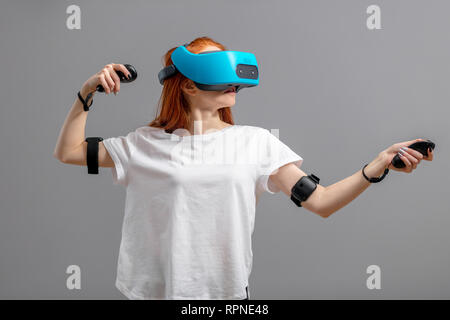 Femme rousse en t-shirt blanc portant l'aide du casque de réalité virtuelle, l'expérience tout en jouant des jeux vidéo, sur fond gris isolé Banque D'Images