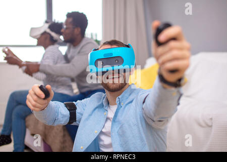 Cheerful young guy sentiment d'excitation et de joie uexperiencing avec casque de réalité virtuelle vr pendant que ses amis africains l'apprentissage de l'utilisation d'un nouveau périphérique Banque D'Images