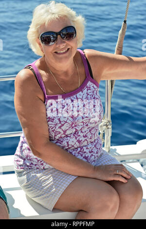 Close-up portrait of smiling mature woman in sunglasses qui est assis sur le pont du bateau de loisirs près de la main courante en métal brillant sur fond de l'eau bleu. Banque D'Images