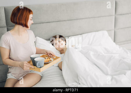 Jolie jeune fille apportant le petit-déjeuner à sa sœur couchée sur le lit. La photo en gros. copie espace, l'amour, l'amitié Banque D'Images