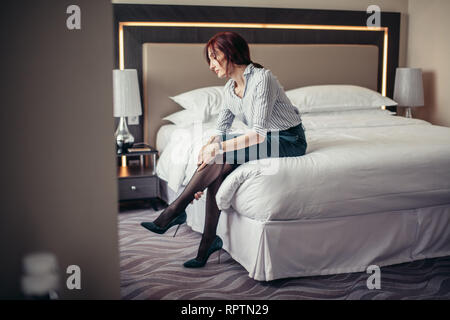 Red-haired dame élégante toucher avec les bras les jambes, être fatigué de porter des talons alors qu'il était assis dans la chambre une fois de retour à la maison de l'office. Banque D'Images