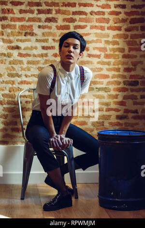 Belle jeune femme, modèle de la mode, avec très peu de coupe, looking at camera assis sur une chaise, avec un mur de briques à l'arrière-plan. Banque D'Images