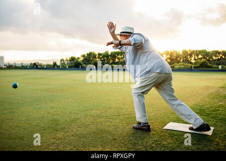 Vieil homme jouant un jeu de boules. Old man in hat jette une boules en position debout dans une pelouse. Banque D'Images