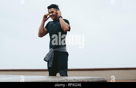 Fit young asian man in sportswear wearing casque à écouter de la musique à l'extérieur lors de la formation. Smiling sportsman prenant pause dans la formation et listenin Banque D'Images