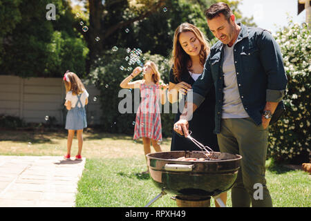 Heureux l'homme et de la femme en se tenant ensemble barbecue dans leur cour. Les filles jouant avec des bulles de savon pendant que leurs parents faire cuire la nourriture pour l'occasion Banque D'Images