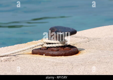Vieux fer rouillé partiellement forte amarre bollard vissé sur la jetée en pierre locale avec cinq vis de métal rouillé avec blanc corde autour de Banque D'Images