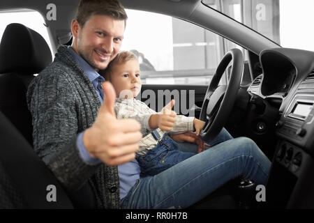 Père et fils assis dans le siège du conducteur de voiture chère nouvelle, showing Thumbs up. Parent et enfant posant en location de chalet, smiling, looking at camera, boy holding main sur le volant. Banque D'Images