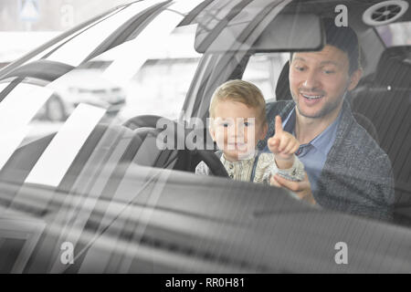 Vue avant du pare-brise par l'intermédiaire du père et fils assis dans le siège du conducteur de l'automobile et l'observation d'Location de cabine. Peu cute boy looking at camera, en pointant avec le doigt. Banque D'Images