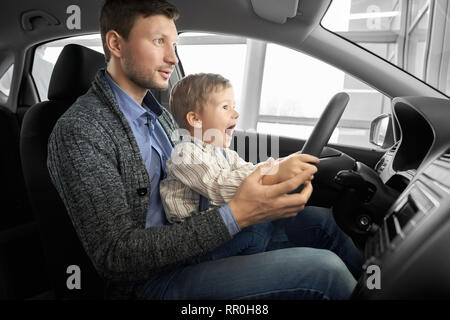 Surprise garçon avec bouche ouverte assis sur les genoux du père dans la nouvelle voiture. Parent et fils assis dans le siège du conducteur, tenant les mains sur le volant, l'essai et l'observation d'automobile. Banque D'Images