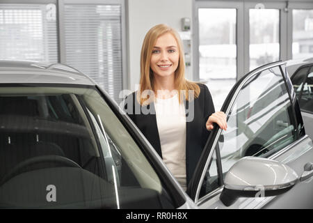 Belle femme aux cheveux blonds, debout près de cher automobile avec porte ouverte. Femme manager de l'agence de voiture posant en showroom, souriant et regardant la caméra. Banque D'Images