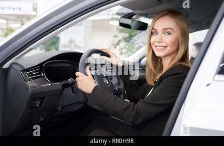 Votre femelle sitting in car cabine de nouveau, cher automobile. Belle blonde woman holding hands au volant, à la caméra et au sourire. Manager travaillant en concession. Banque D'Images