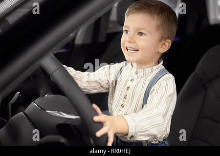 Mignon, joli petit garçon assis dans le siège conducteur de voiture cabine. Heureux l'enfant se tenant la main sur le volant, à la voiture, en souriant. Concept de voiture d'exposition. Banque D'Images