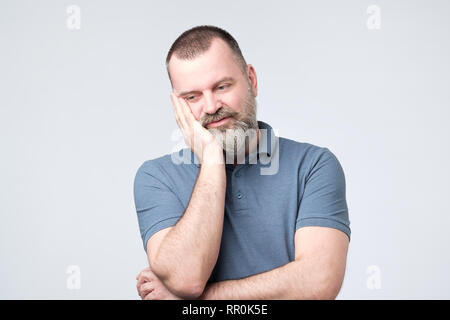 Ennuyer homme adulte avec barbe tenant la main sur la joue alors que le soutien à l'aide d'un autre main croisés Banque D'Images