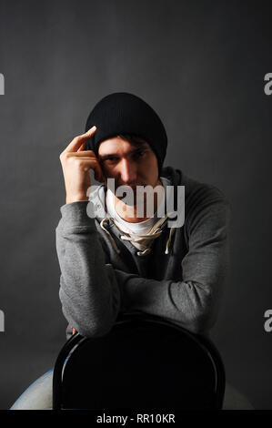 Le jeune homme assis sur une chaise dans un sweat gris et un chapeau noir tenant sa main à sa tête à l'ombre. Banque D'Images