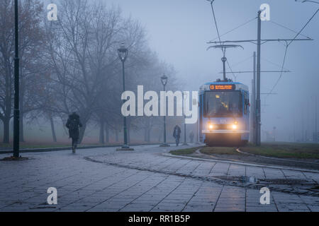 Crépuscule sur un hiver froid et humide journée. Train électrique avec des projecteurs et des gens qui marchent. Les arbres dénudés et les lampes de rue sont sur le chemin. Banque D'Images