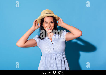 Freestyle. Femme en robe et hat standing isolé sur mur bleu à la joyeuse de l'appareil photo Banque D'Images