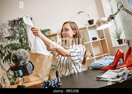 Fashion blogger. Heureux et cute woman showing chemise blanche sur l'appareil-photo pendant l'enregistrement d'une nouvelle vidéo pour son blog de mode. La beauté. Video review. Blogging Banque D'Images
