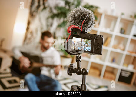 Apprentissage de la guitare. Musique mâle blogger assis sur le plancher à la maison et l'enregistrement de nouveaux cours de guitare avec l'appareil photo sur le premier plan, la création de contenu. Youtube Banque D'Images